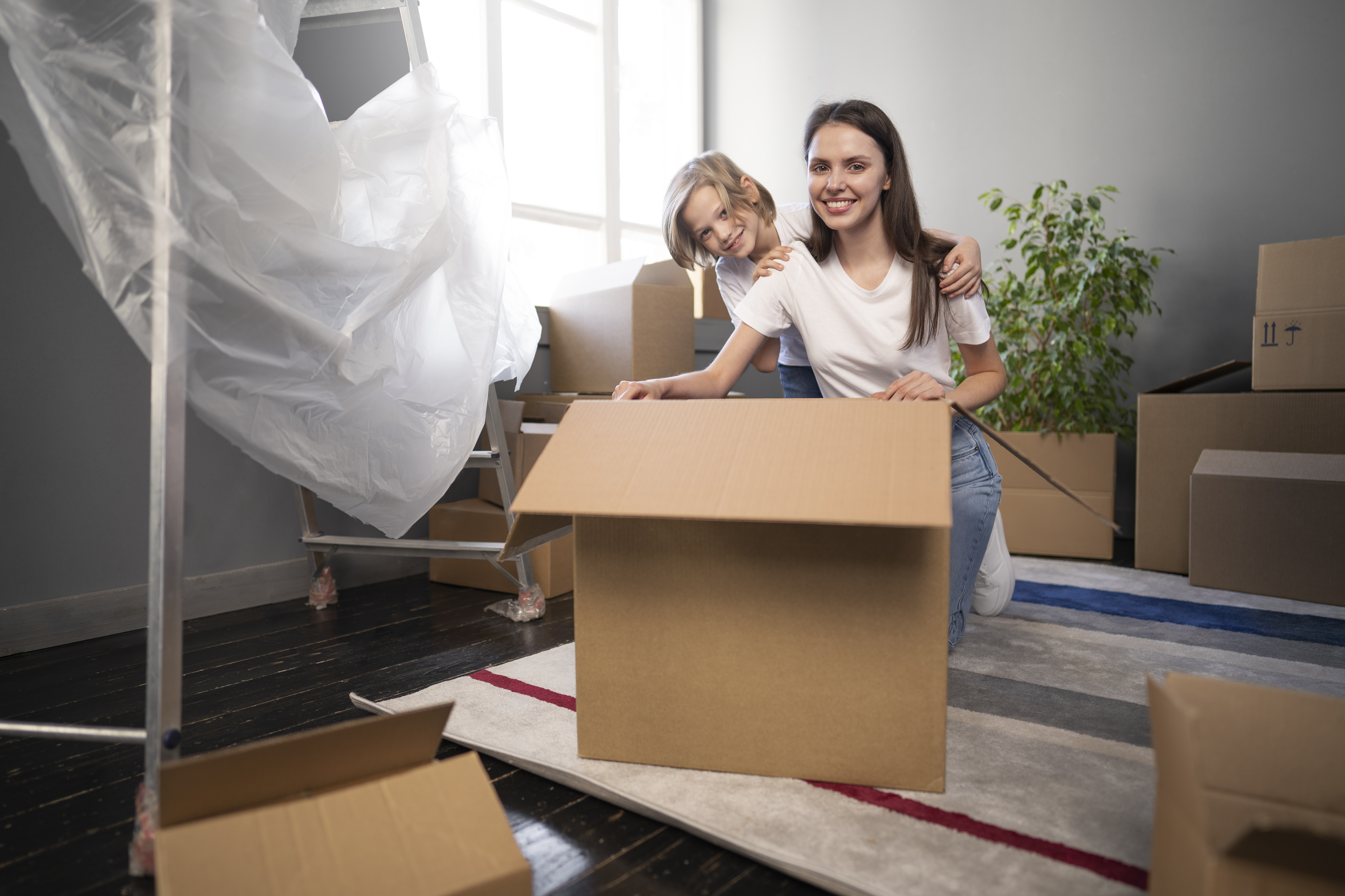 Photo of a young woman and young child smiling at the camera. They are kneeling behind a large cardboard box and surrounded by other boxes