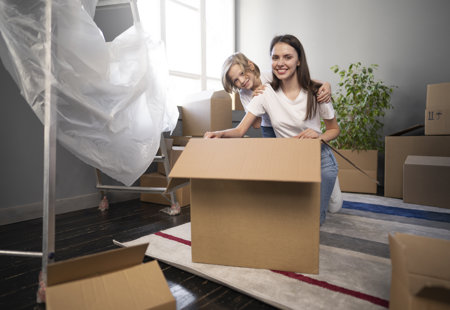 Photo of a young woman and young child smiling at the camera. They are kneeling behind a large cardboard box and surrounded by other boxes