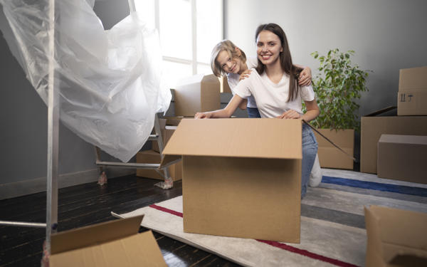 Photo of a young woman and young child smiling at the camera. They are kneeling behind a large cardboard box and surrounded by other boxes