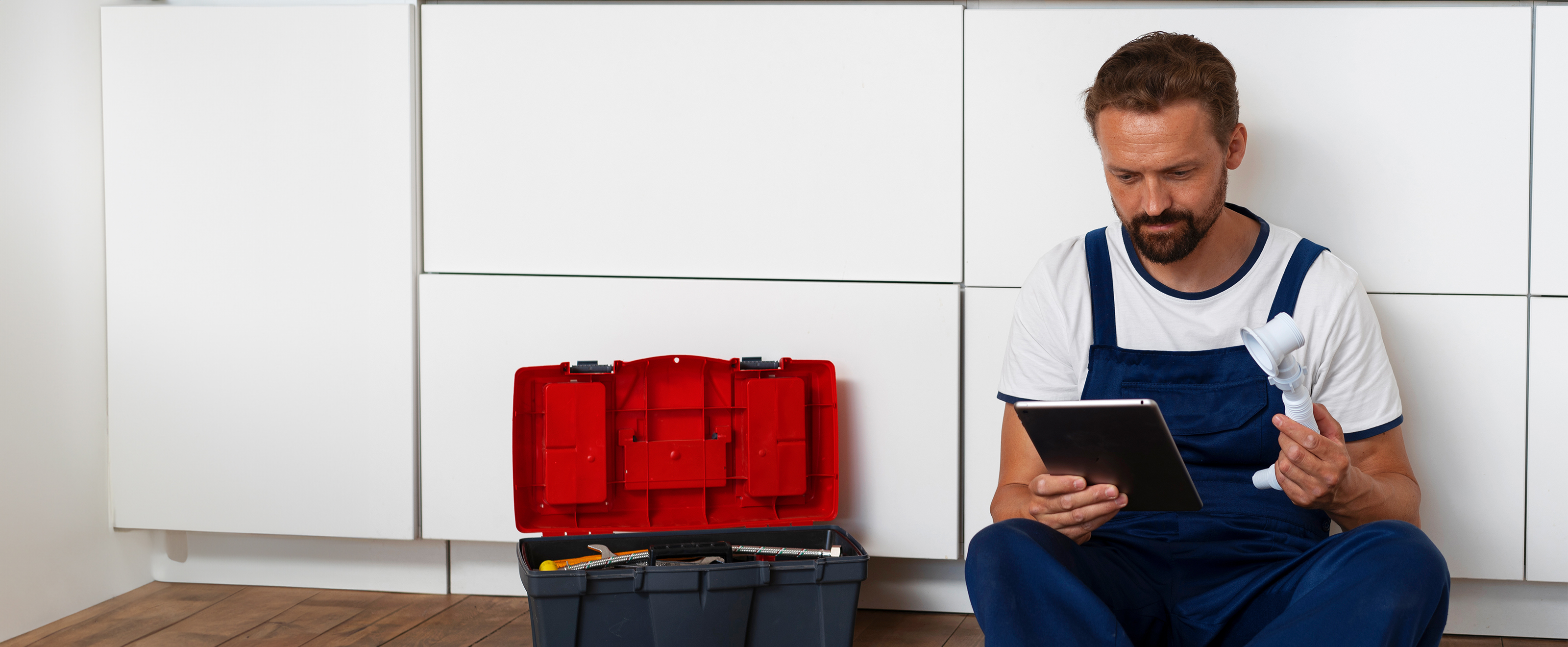 Photo of a man wearing overalls sitting on the floor in front of kitchen cabinets. He is using a tablet computer and there is a toolbox to the side of him.
