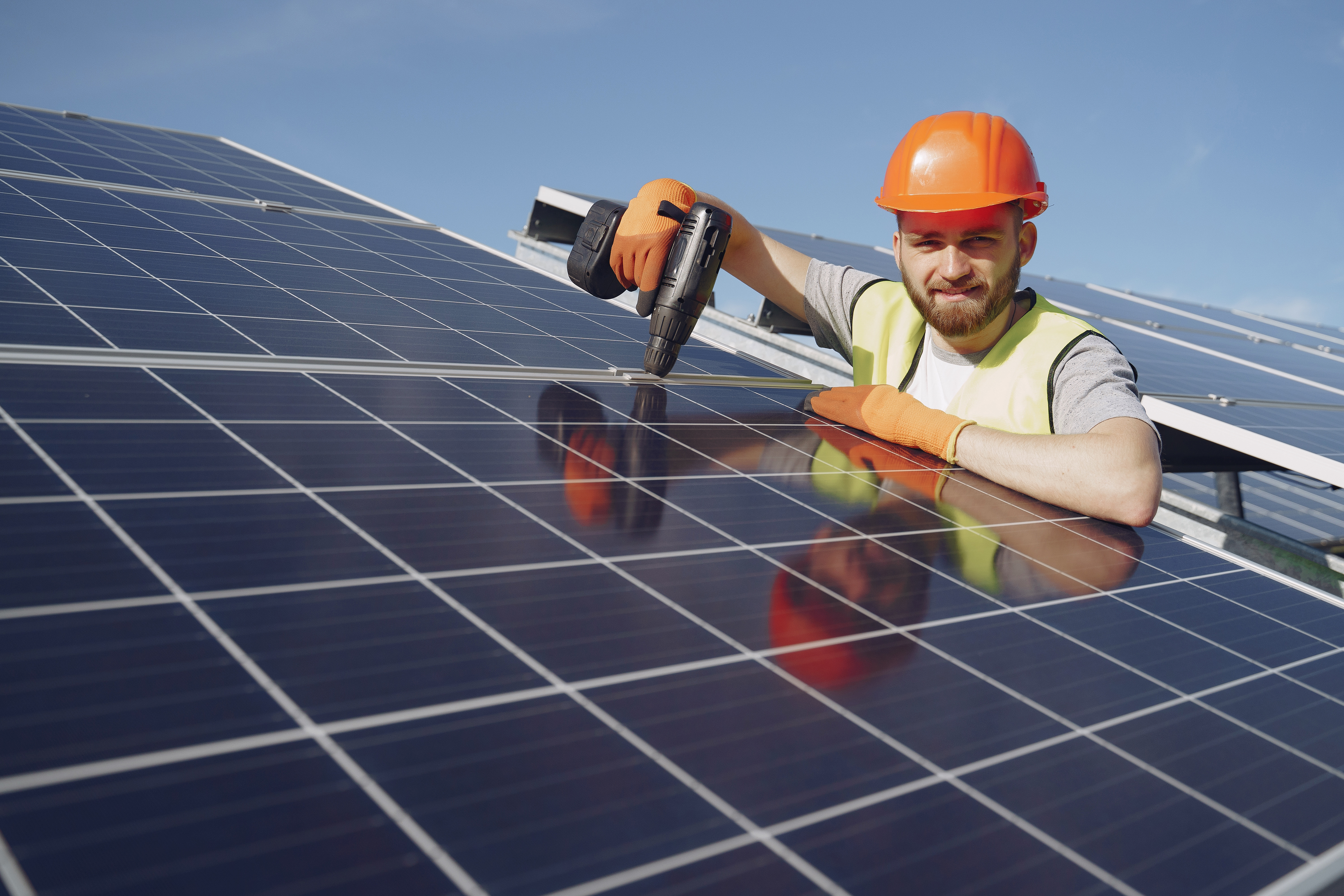 Photo of a workman in a hard hat holding a drill and installing electrical solar panels on a roof
