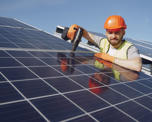 Photo of a workman in a hard hat holding a drill and installing electrical solar panels on a roof