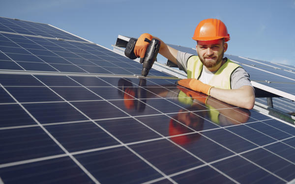 Photo of a workman in a hard hat holding a drill and installing electrical solar panels on a roof