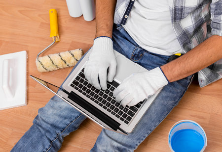 Photo take from above of a man sitting on the floor. He is using a laptop that is lying over his legs. There are various decorating and DIY tools on the floor around him. 