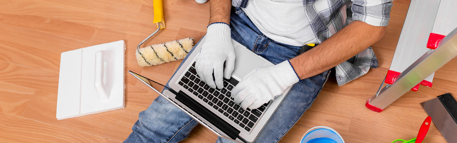 Photo take from above of a man sitting on the floor. He is using a laptop that is lying over his legs. There are various decorating and DIY tools on the floor around him. 