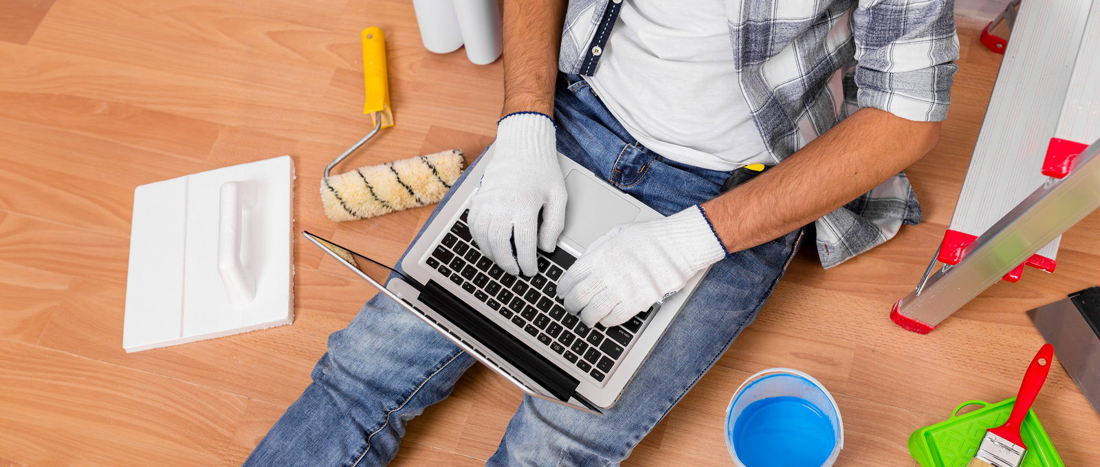 Photo take from above of a man sitting on the floor. He is using a laptop that is lying over his legs. There are various decorating and DIY tools on the floor around him. 