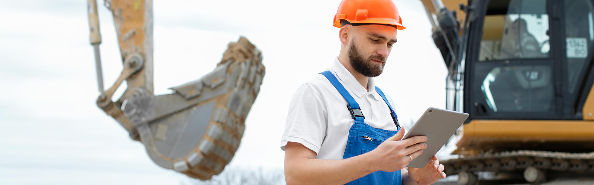 Photo of a construction worker in a hard hat and holding a clipboard. He is standing in front of an excavator