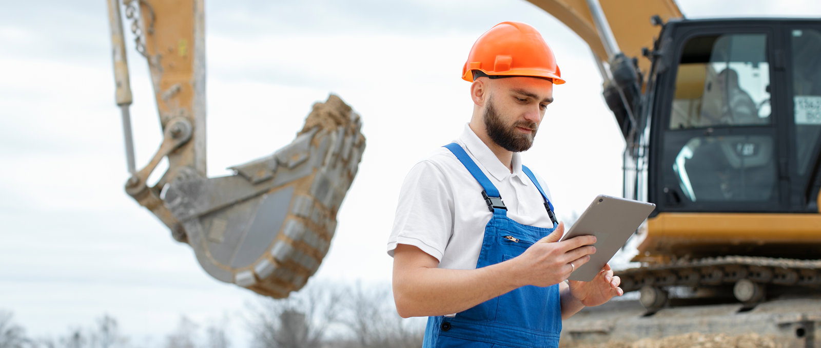 Photo of a construction worker in a hard hat and holding a clipboard. He is standing in front of an excavator