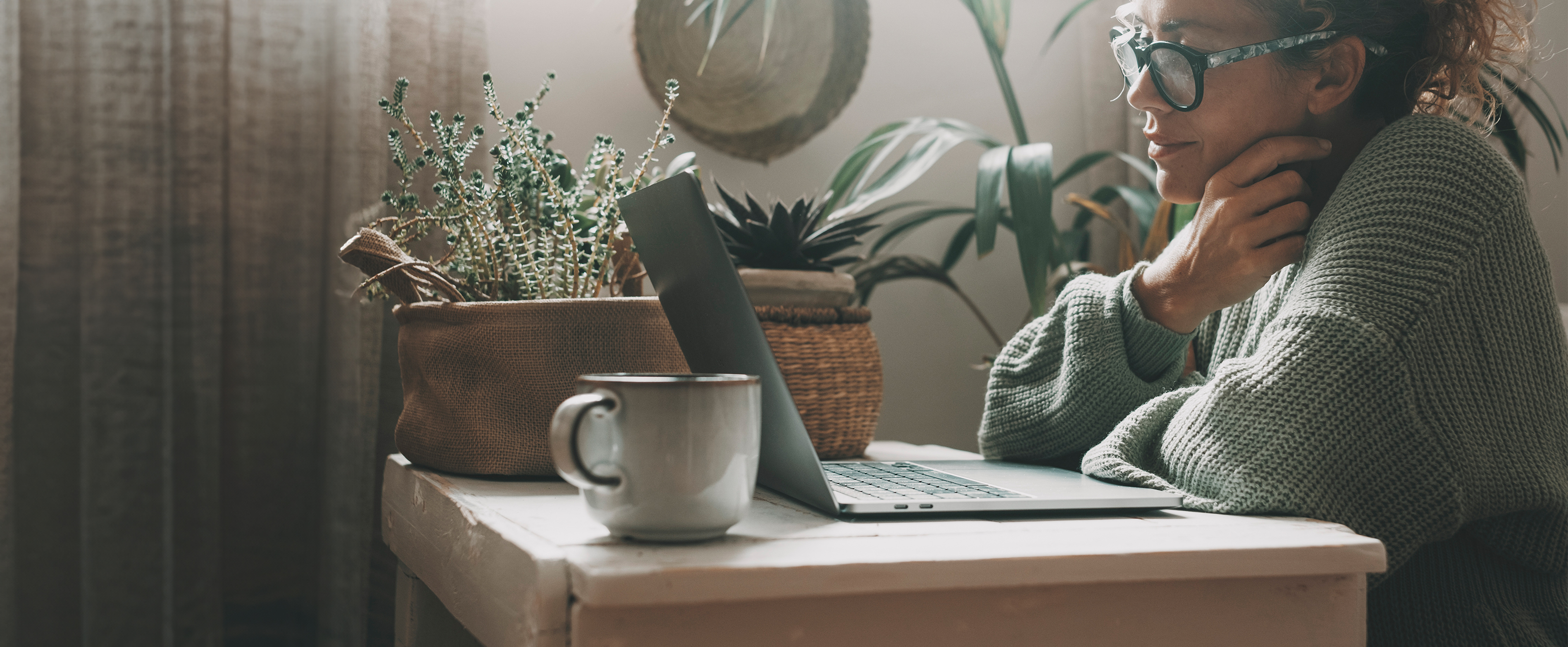Photo of a woman sitting at a table looking at a laptop. The setting is relaxed, probably at home rather than in an office