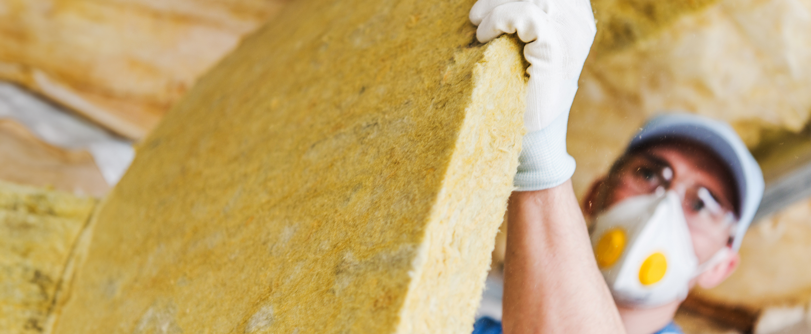 Photo of a man in a roof space wearing a protective mask holding a large sheet of insulation material towards the camera