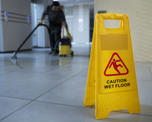 Close up photo of a 'caution - wet floor' sign standing on a tiled floor. Someone is cleaning the floor in the background.