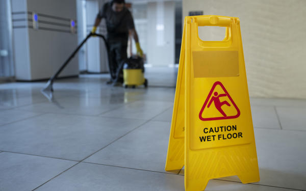 Close up photo of a 'caution - wet floor' sign standing on a tiled floor. Someone is cleaning the floor in the background.