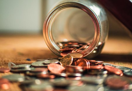 close up photo of a glass jar lying overturned on a surface with lots of coins spilling out of it onto the surface