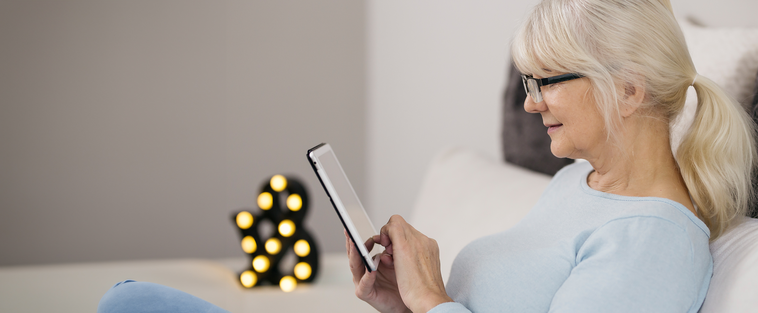 Photo of an older woman wearing glasses sitting on a sofa and using a tablet computer