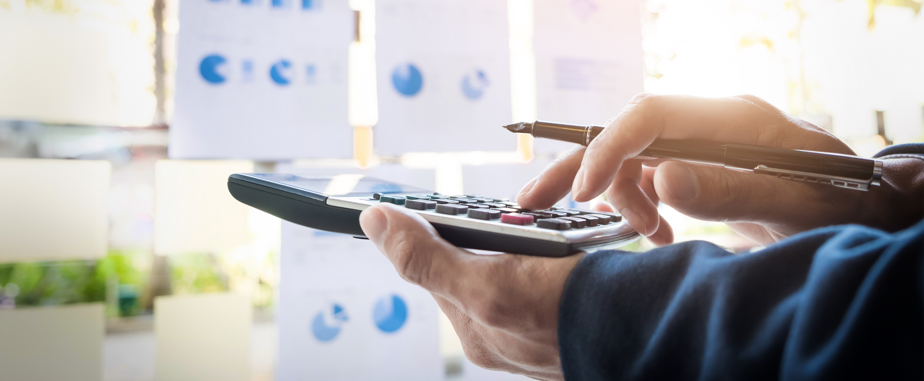 close-up photo of someone's hands holding a large calculator. In the background are various sheets of paper with graphs and charts on. 