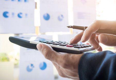 close-up photo of someone's hands holding a large calculator. In the background are various sheets of paper with graphs and charts on. 