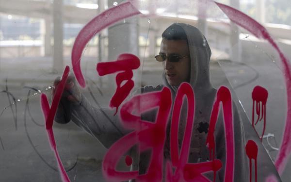 Photo of a man in sunglasses and a hoodie spraying red graffiti on glass - viewed from the other side of the glass