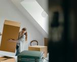 Photo of a woman lifting a large carboard box in what looks like a bedroom. There are other boxes and a suitcase visible