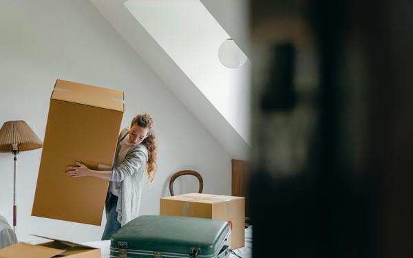 Photo of a woman lifting a large carboard box in what looks like a bedroom. There are other boxes and a suitcase visible