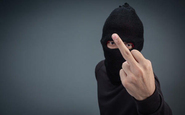 Photo of a man with his face partially covered by black facemask and he is raising a finger towards the camera in a threatening way