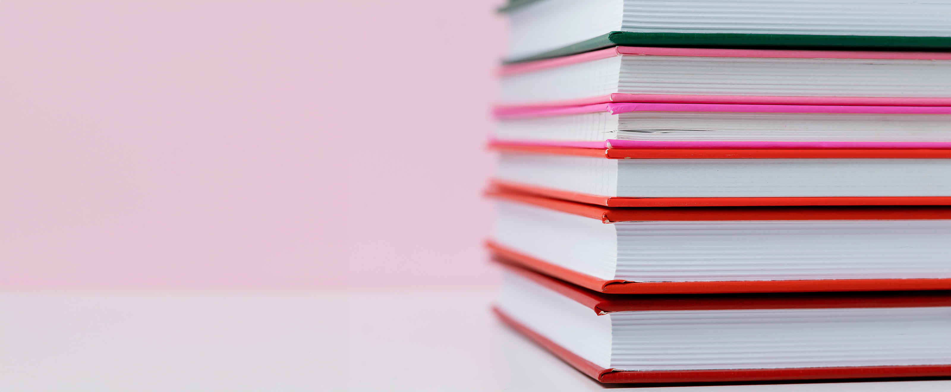 Photo of a stack of multicoloured books on a pink and white background. The books are on the right side of the image and go out of frame.