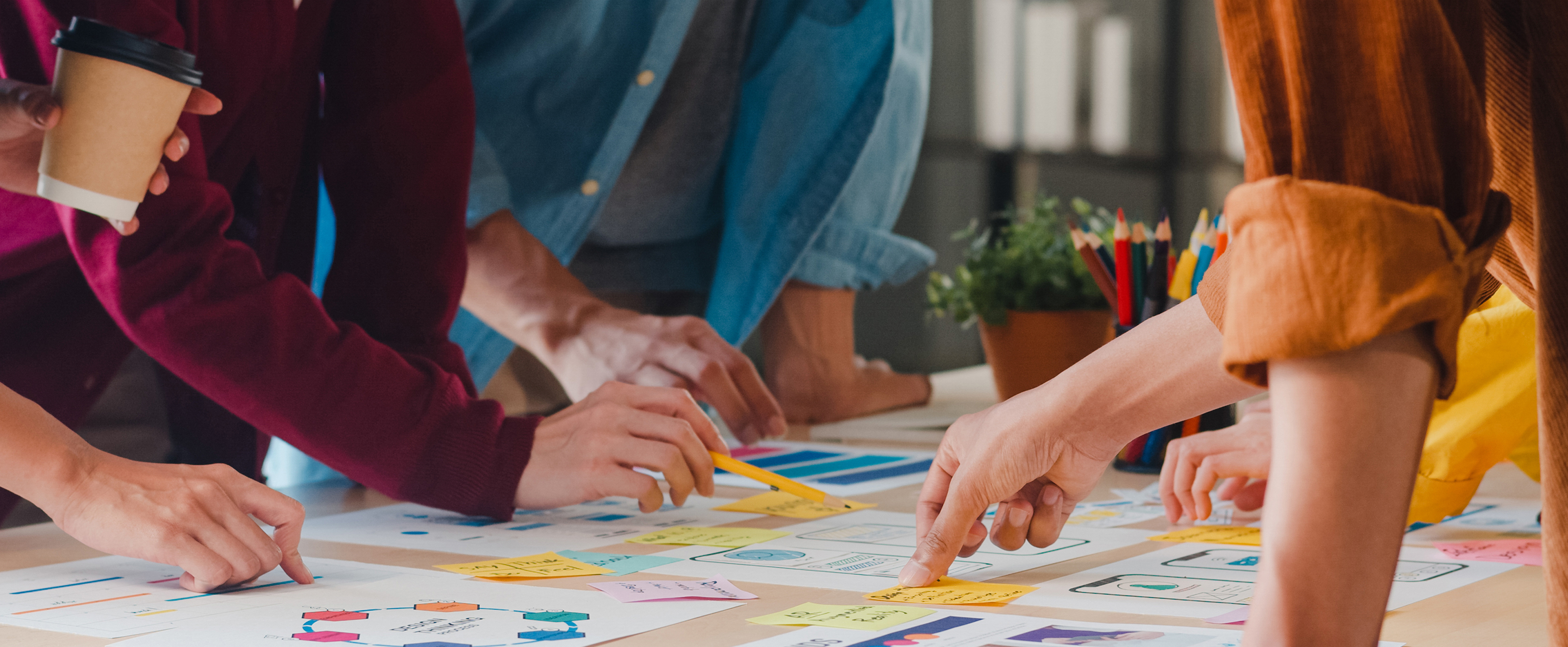 Close-up photo of a table top. There are lots of documents and charts on the table. Several people are standing around pointing at some of the items on the table