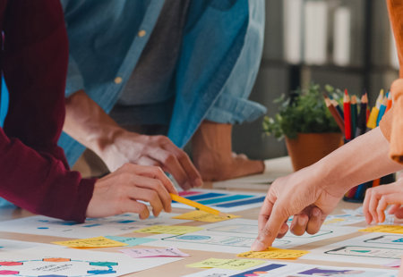 Close-up photo of a table top. There are lots of documents and charts on the table. Several people are standing around pointing at some of the items on the table