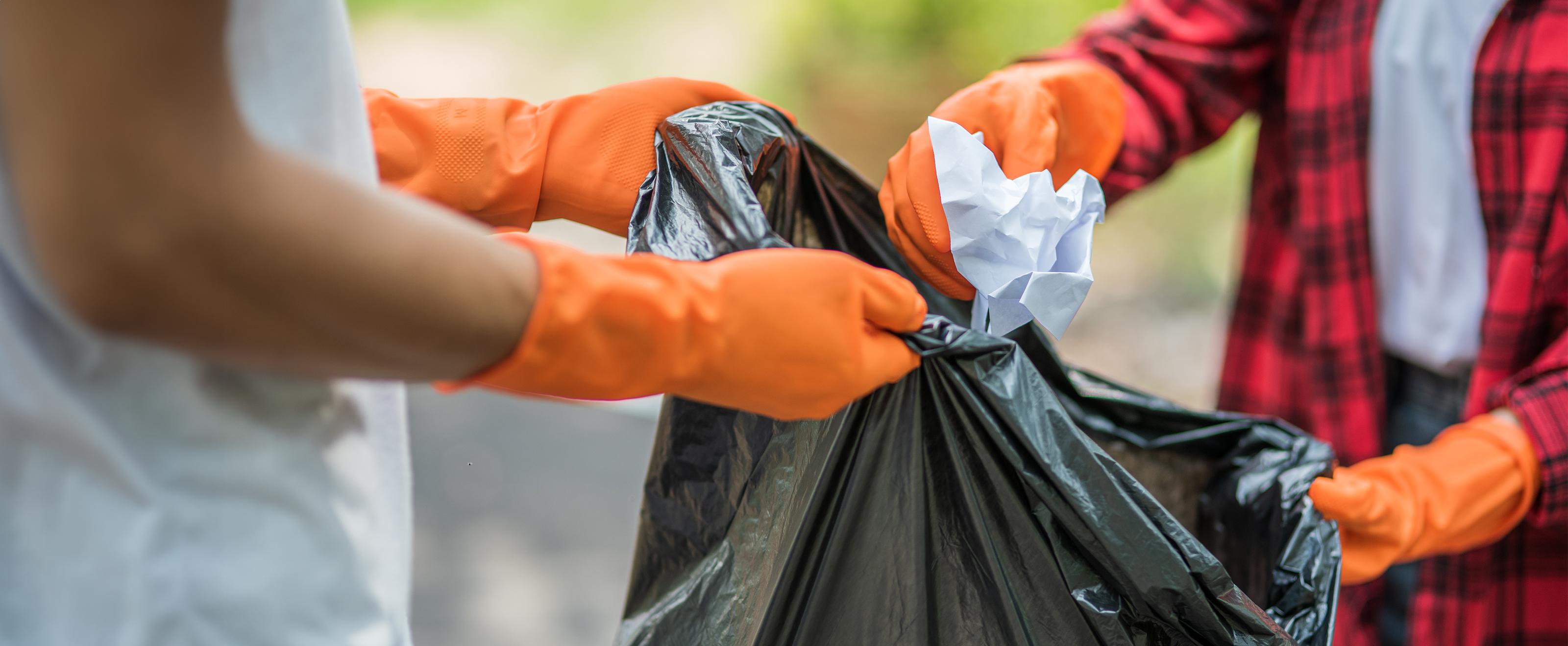 close up photo of someone holding open a black plastic bag. Another person is putting rubbish in the bag. Both people are wearing orange plastic gloves.