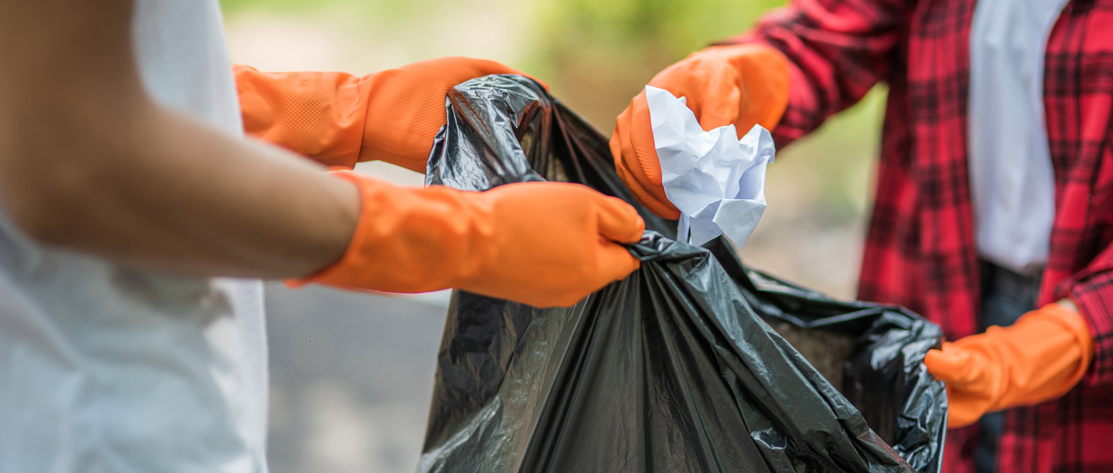 close up photo of someone holding open a black plastic bag. Another person is putting rubbish in the bag. Both people are wearing orange plastic gloves.